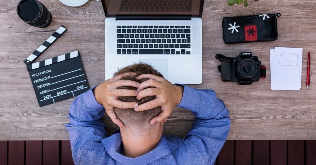 man sitting at a desk with a laptop, film camera, and notes. He is clutching his head in frustration as a representative of content creator burnout