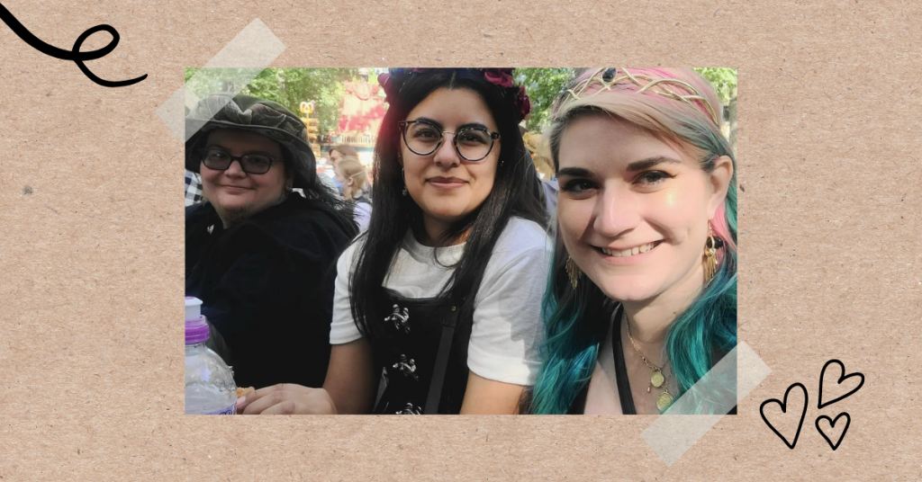 photo that looks like it has been taped to a corkboard. the photo has 3 people, one person (Acorn) on the left wearing glasses and a hat with a feather, a young woman (Bri) in the middle wearing glasses, a flower crown, and a black corset, another woman (Glo) on the right with pink and blue hair wearing a golden tiara